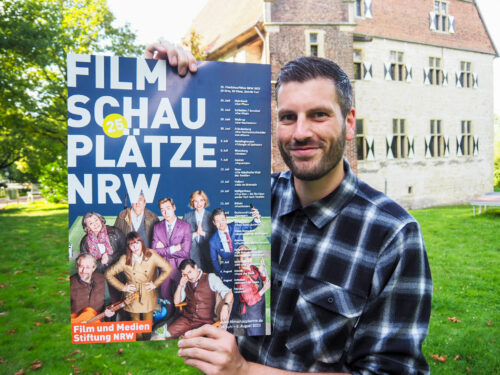 Lukas Bertels von der Kolvenburg zeigt das Plakat der Filmschauplätze NRW 2023. Am 19. Juli ist mit "Yves Saint Laurent" auch an der Billerbeck Station des Open Air Kinos.